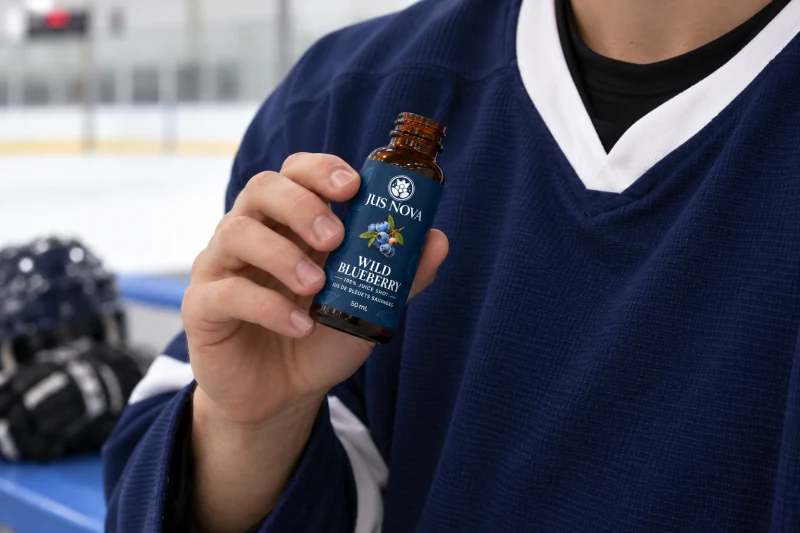 A single hand holding an open Jus Nova Shot 50 mL bottle against a navy hockey jersey, with the face outside the frame and the rink softly blurred behind.