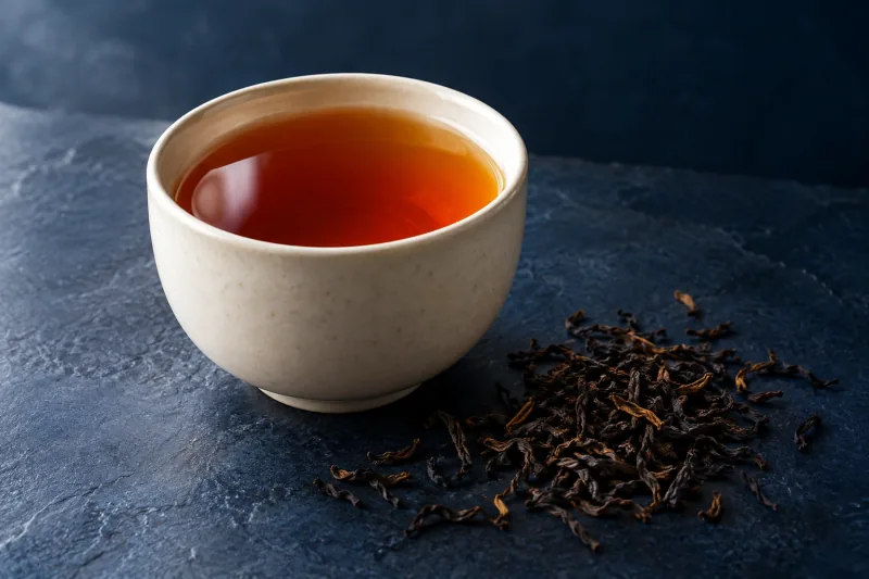Amber tea in a ceramic cup beside loose tea leaves on a navy surface.