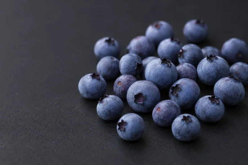 A loose group of small blue-violet wild blueberries with smooth powdery bloom, varied orientations and delicate calyces on a dark studio surface.